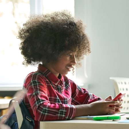 Happy smiling African American teen elementary schoolgirl holding cell phone smartphone sitting in classroom using mobile apps for learning. Modern technologies for education concept. Vertical.